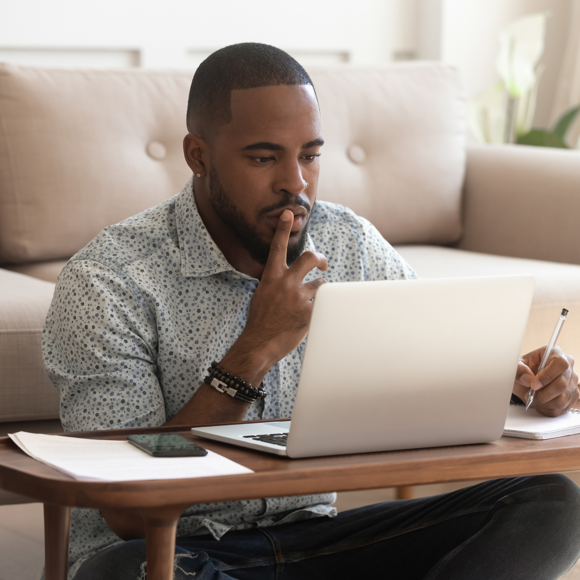 A person sits on the floor, working on a laptop on a low table, while taking notes and appearing to think.