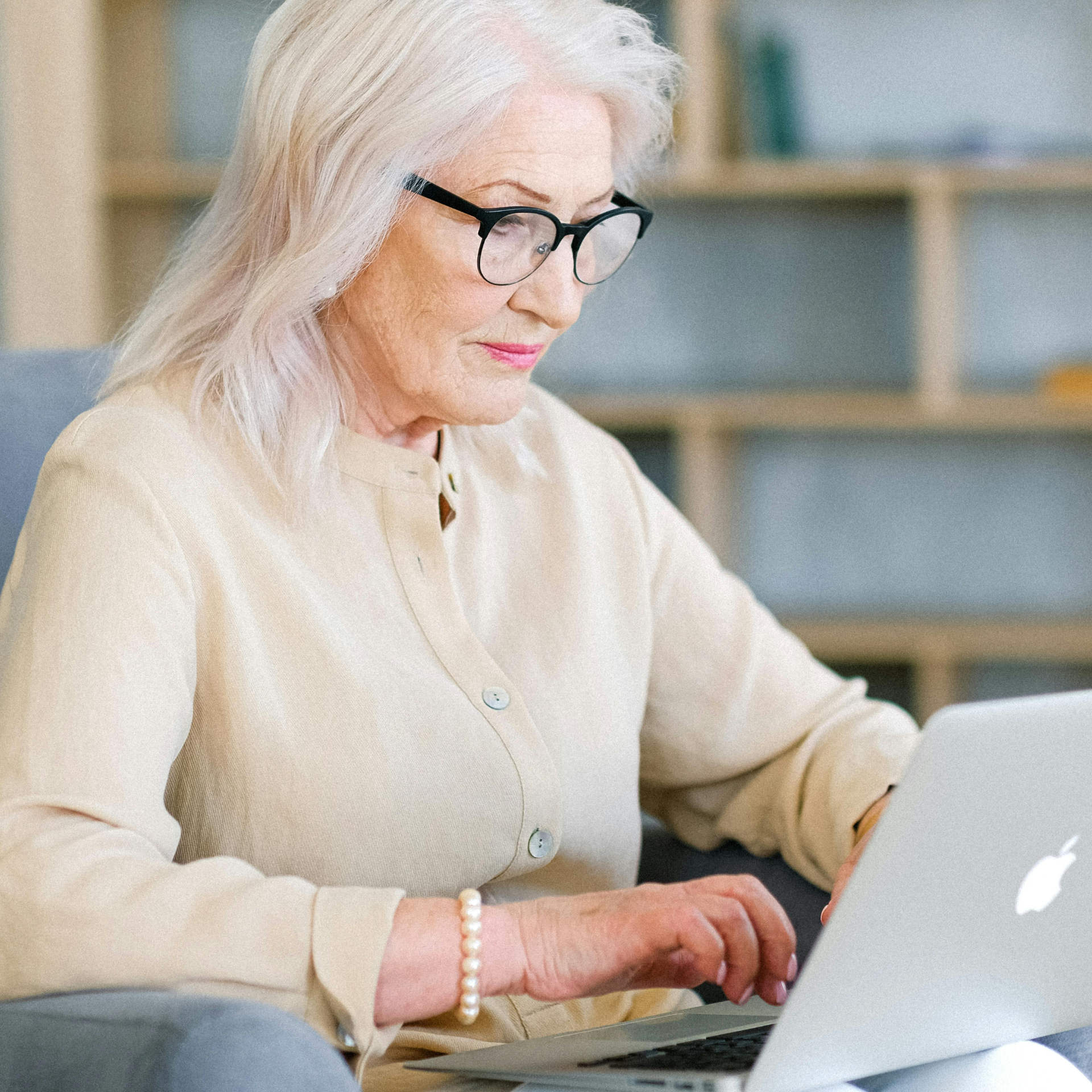 An older woman with glasses uses a laptop, sitting in a chair indoors.