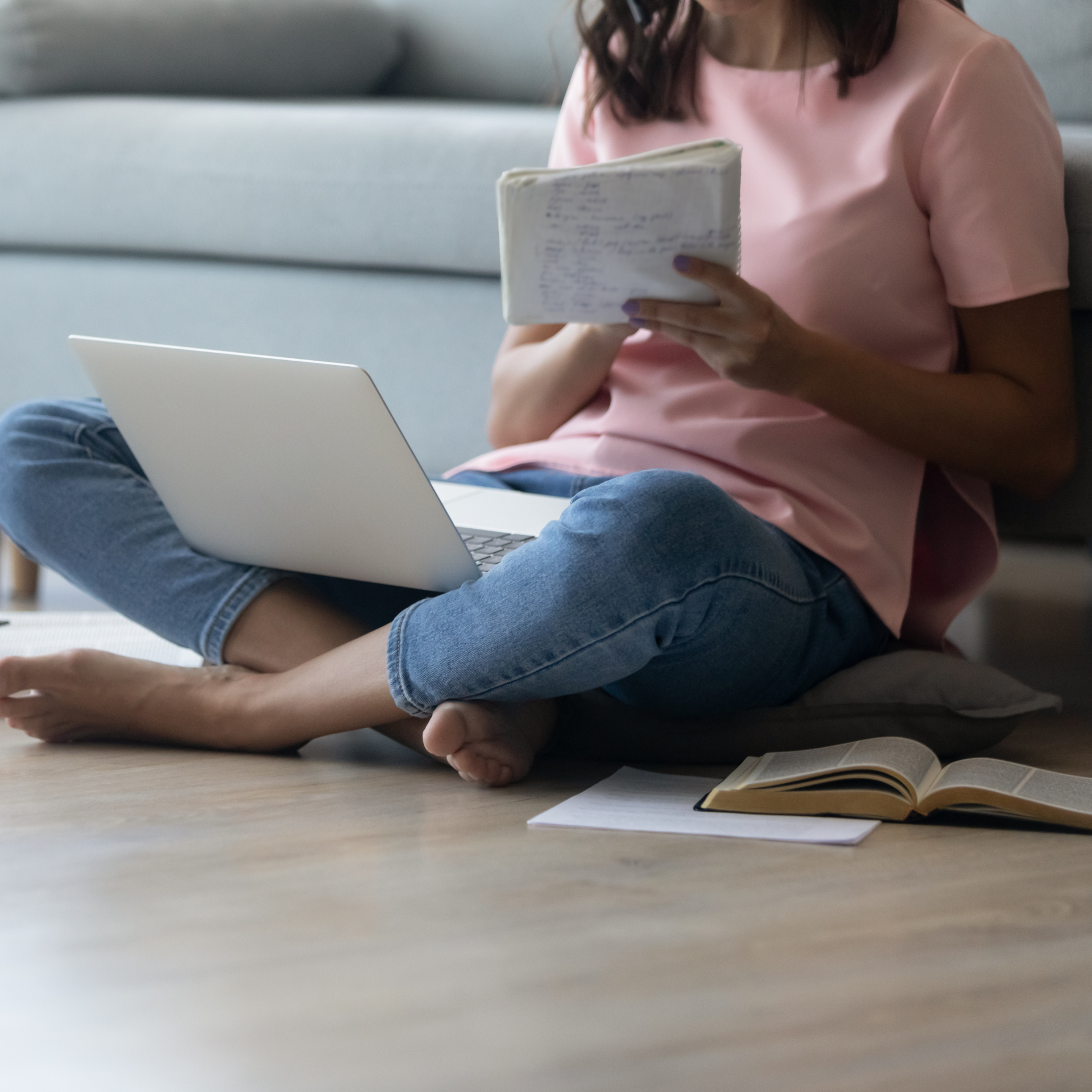 Woman sits cross-legged on floor with laptop, reading book and notes in front of couch.