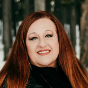 Woman with long red hair smiles, posing outdoors with trees in the background.