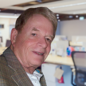 Smiling older man in a brown plaid jacket, white shirt, and light brown hair, in an office setting.