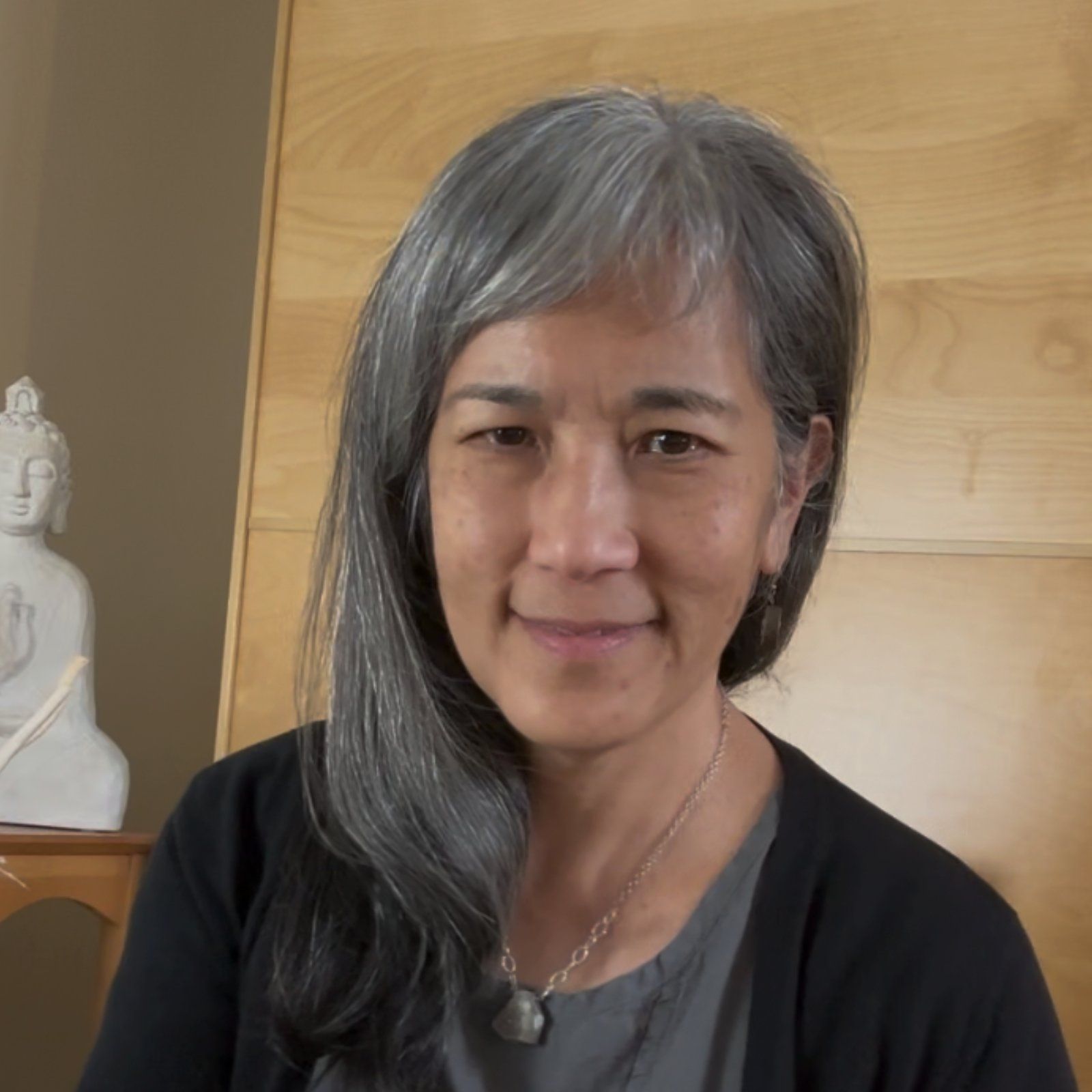 A person with shoulder-length gray hair smiles gently, wearing a dark cardigan and a necklace, with a statue nearby.