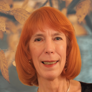 Woman with short red hair smiles against a blurred backdrop of leaves.