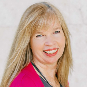 Woman with long blonde hair smiles, wearing a pink blazer and gray shirt.
