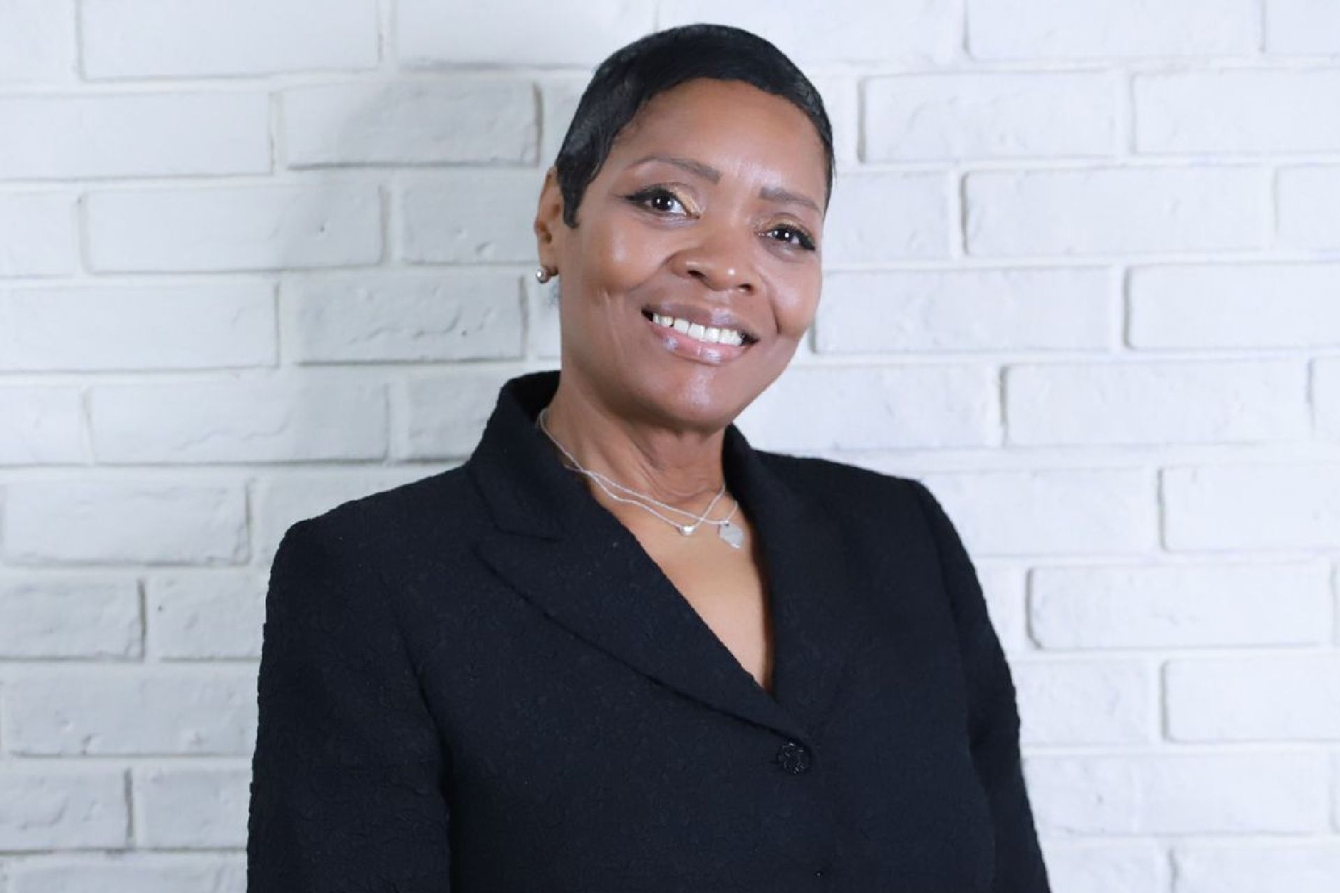 A woman in a black suit is smiling in front of a white brick wall.