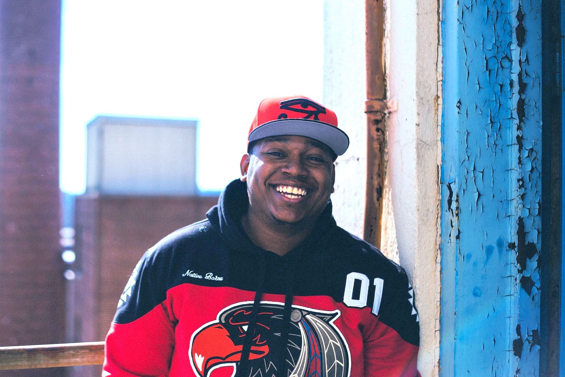 A man wearing a red hoodie and a hat is leaning against a blue wall.