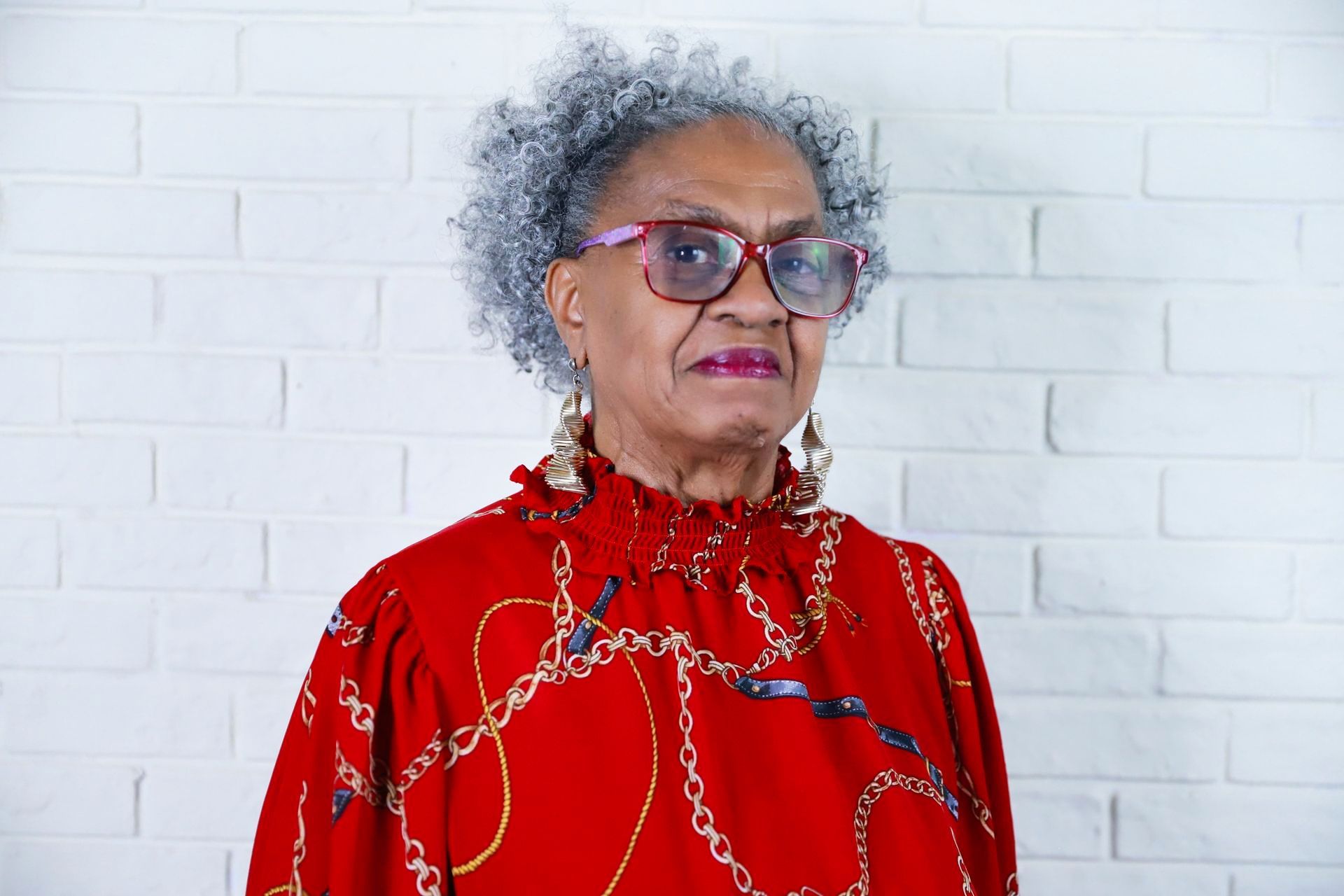 An elderly woman wearing glasses and a red shirt is standing in front of a white brick wall.