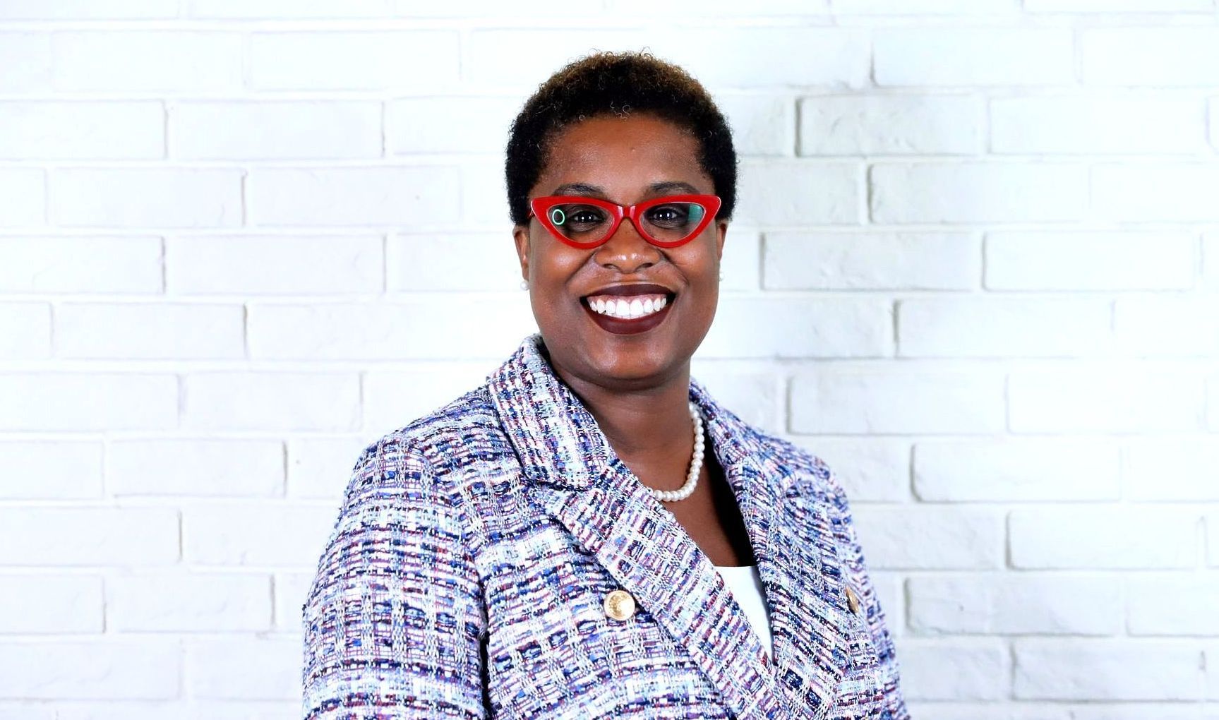 A woman wearing glasses and a jacket is smiling in front of a white brick wall.
