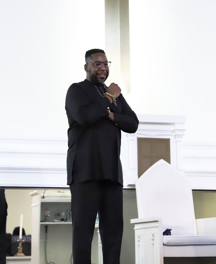 A man in a black suit is standing in front of a white chair