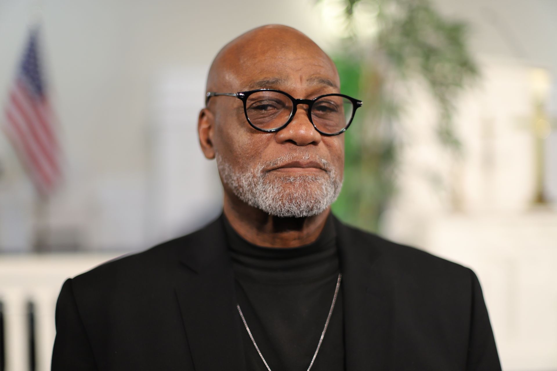A man with glasses and a beard is wearing a black suit and a necklace.