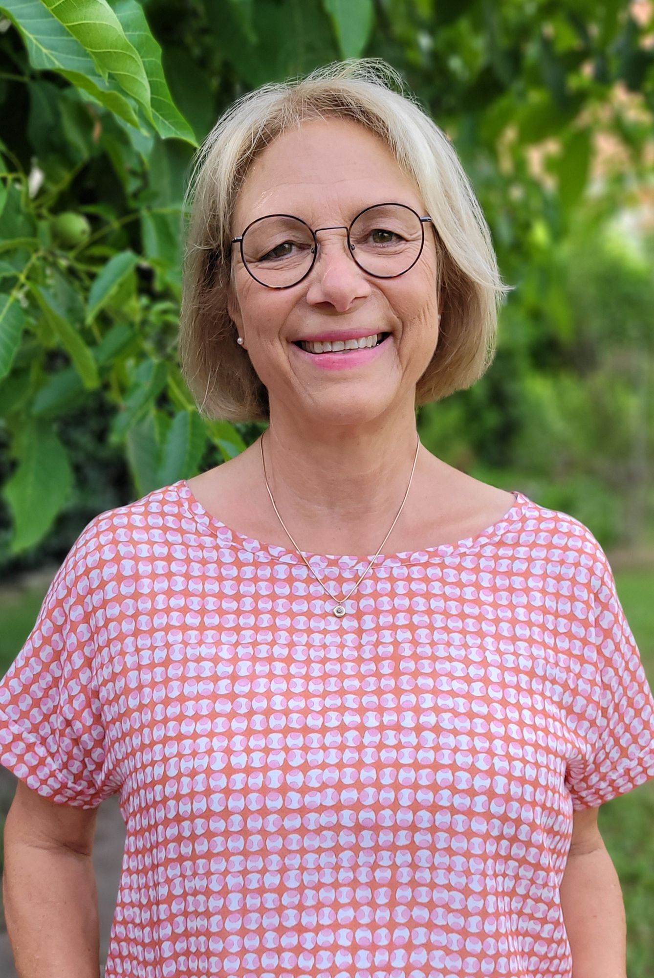 Eine Frau mit Brille und rosa Hemd lächelt vor einem Baum.