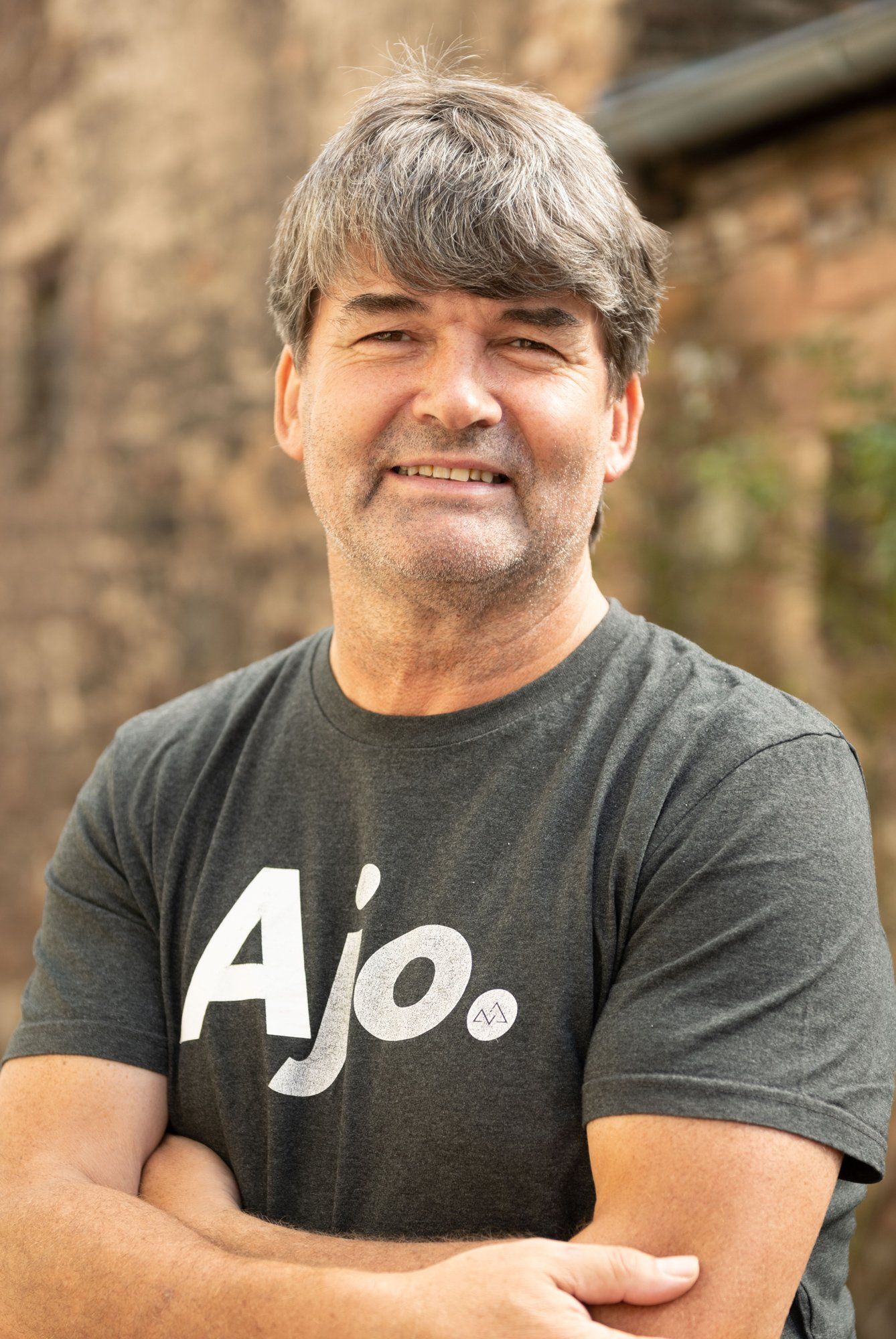 Ein Mann steht mit verschränkten Armen da und trägt ein graues T-Shirt mit der Aufschrift „Ajo“.