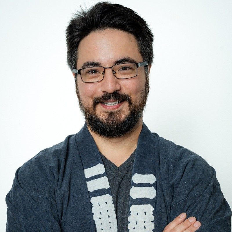A smiling person with glasses and a beard, wearing a dark blue kimono-style jacket, poses with arms crossed against white.