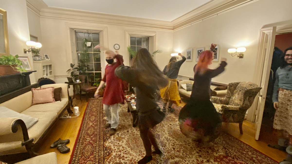 Several people are dancing in a cozy, lamp-lit living room with a large patterned rug and furniture.