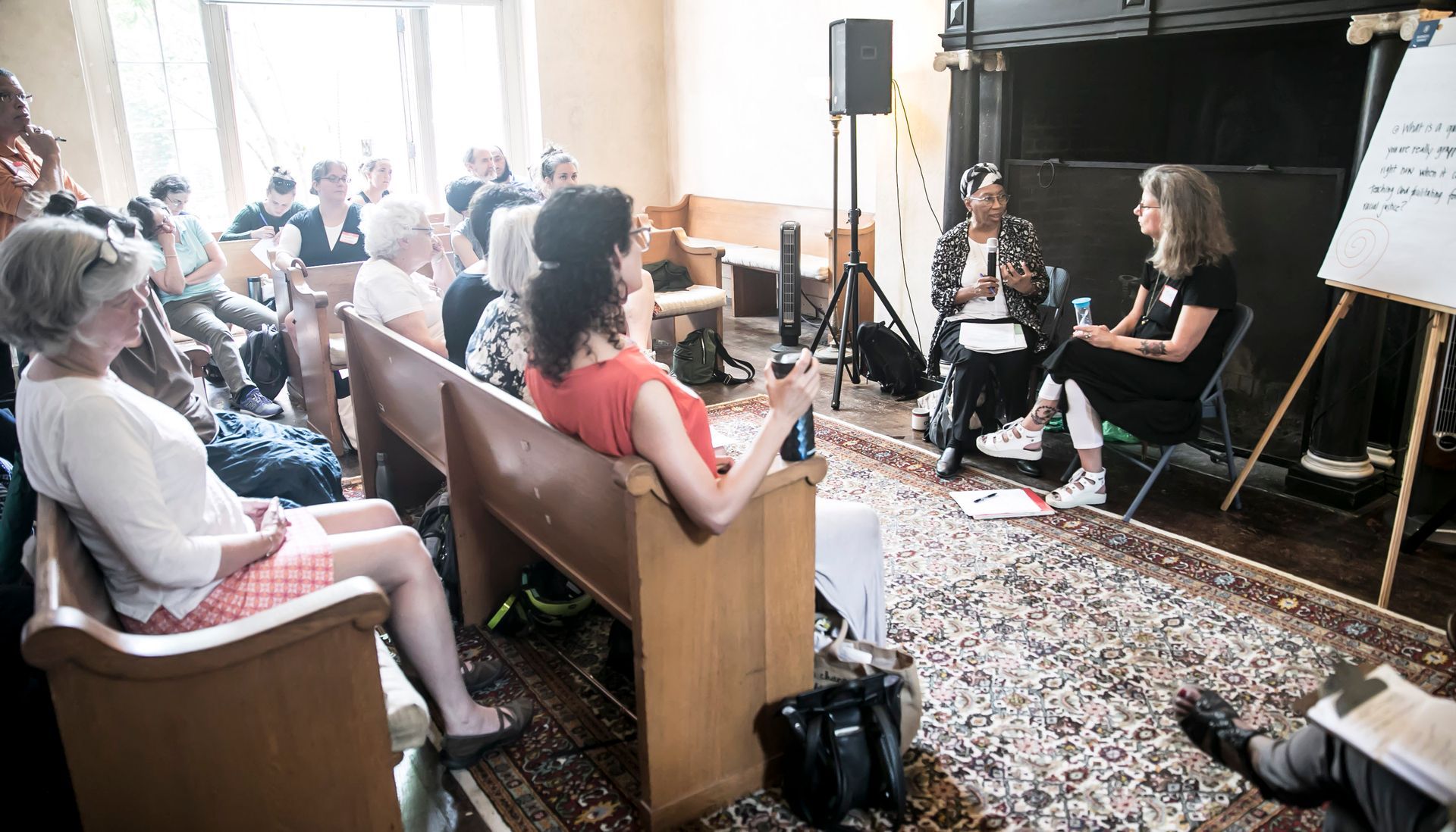 Two speakers lead a discussion for a small, diverse audience seated in wooden pews within an indoor meeting space.