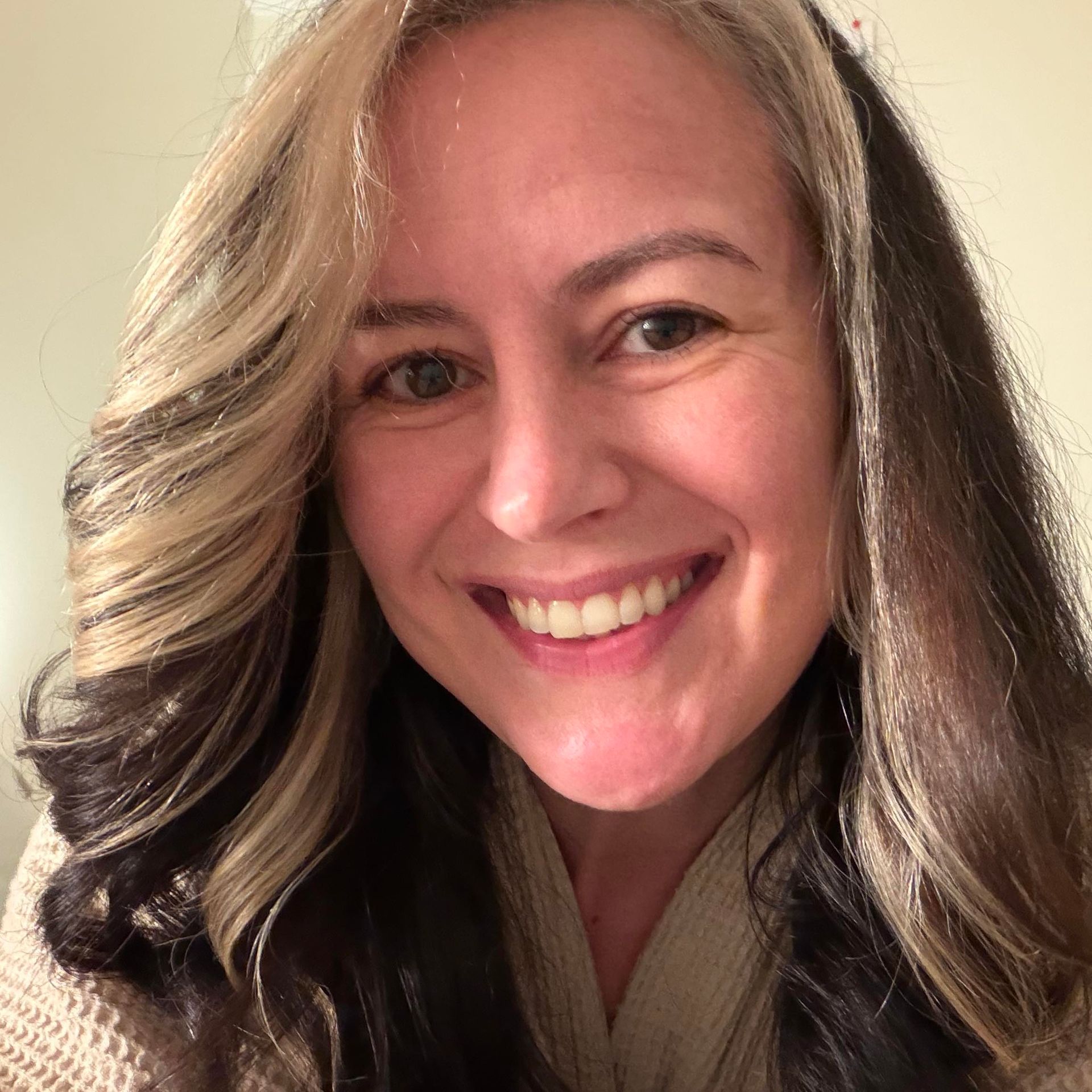 A smiling person with layered, shoulder-length brown hair featuring prominent blonde highlights, wearing a beige top.