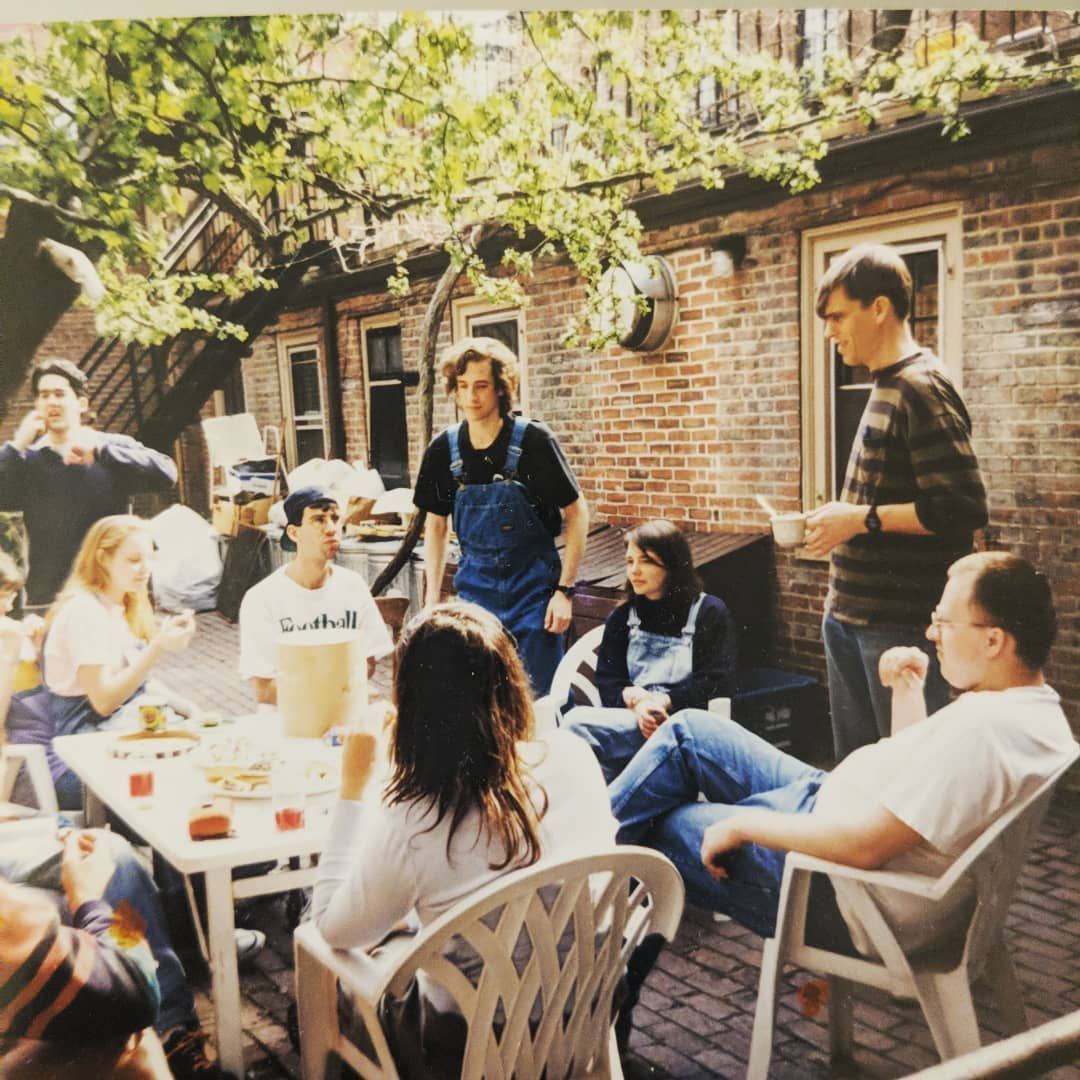 A group of people gathers for an outdoor meal on a brick patio in a residential backyard, seated around a white table.