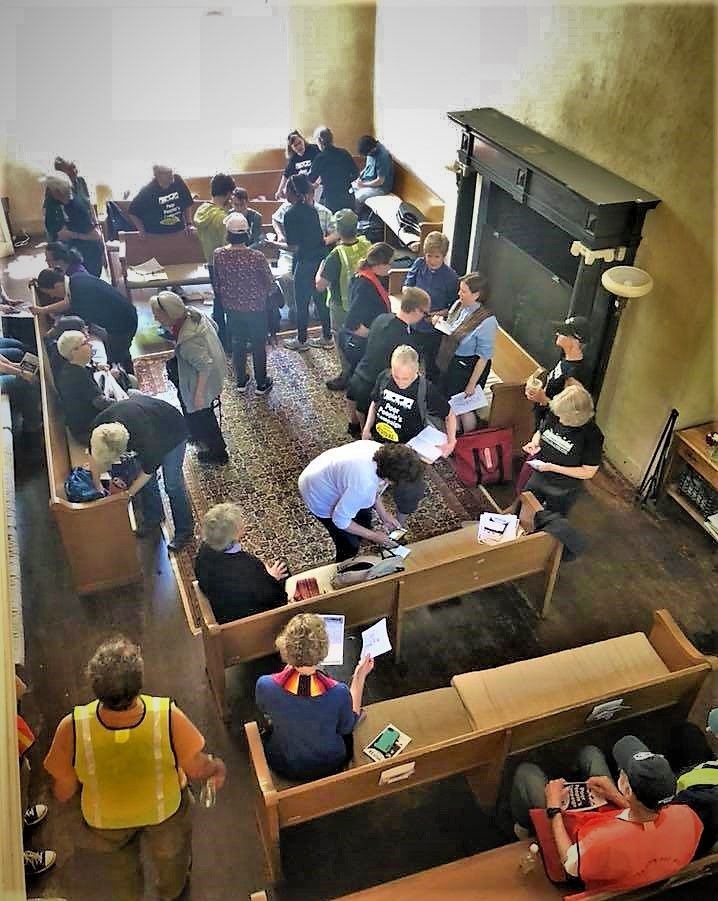 People gather in a room with rows of wooden benches, standing and talking near a large fireplace.
