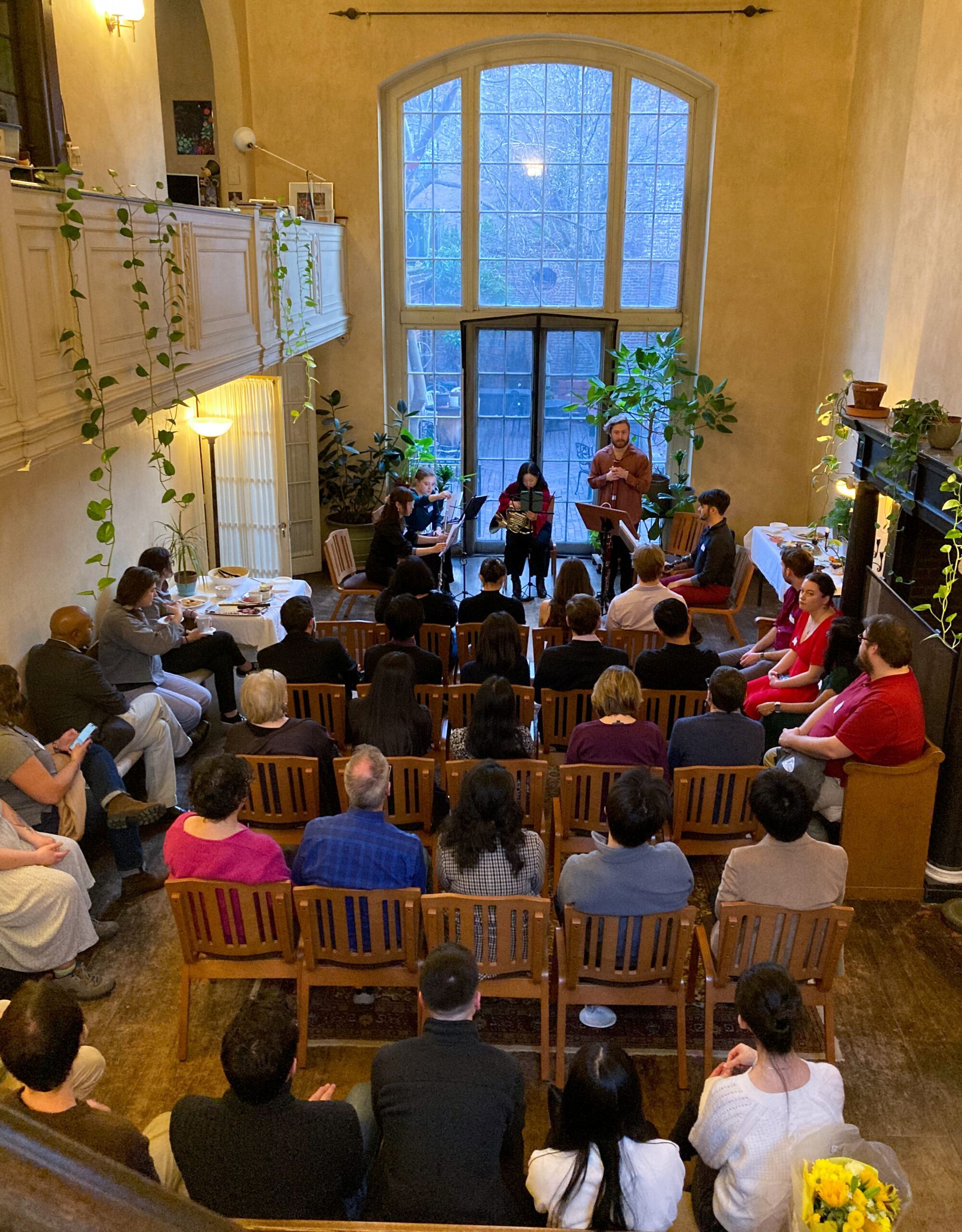 An audience sits in wooden chairs in a hall with tall, arched windows, watching a musical performance on a small stage.