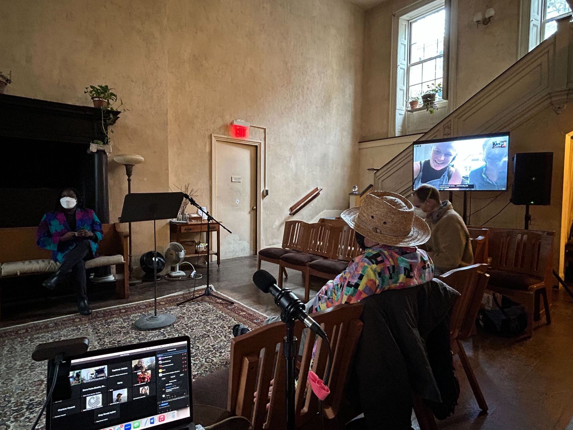 People attend a hybrid meeting in a historic hall with a screen displaying remote participants and a laptop in the front.