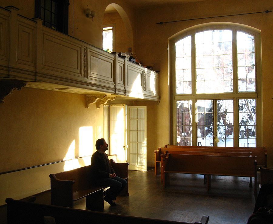 Man sits in a sunlit church.  Wooden pews, balcony, tall arched window.