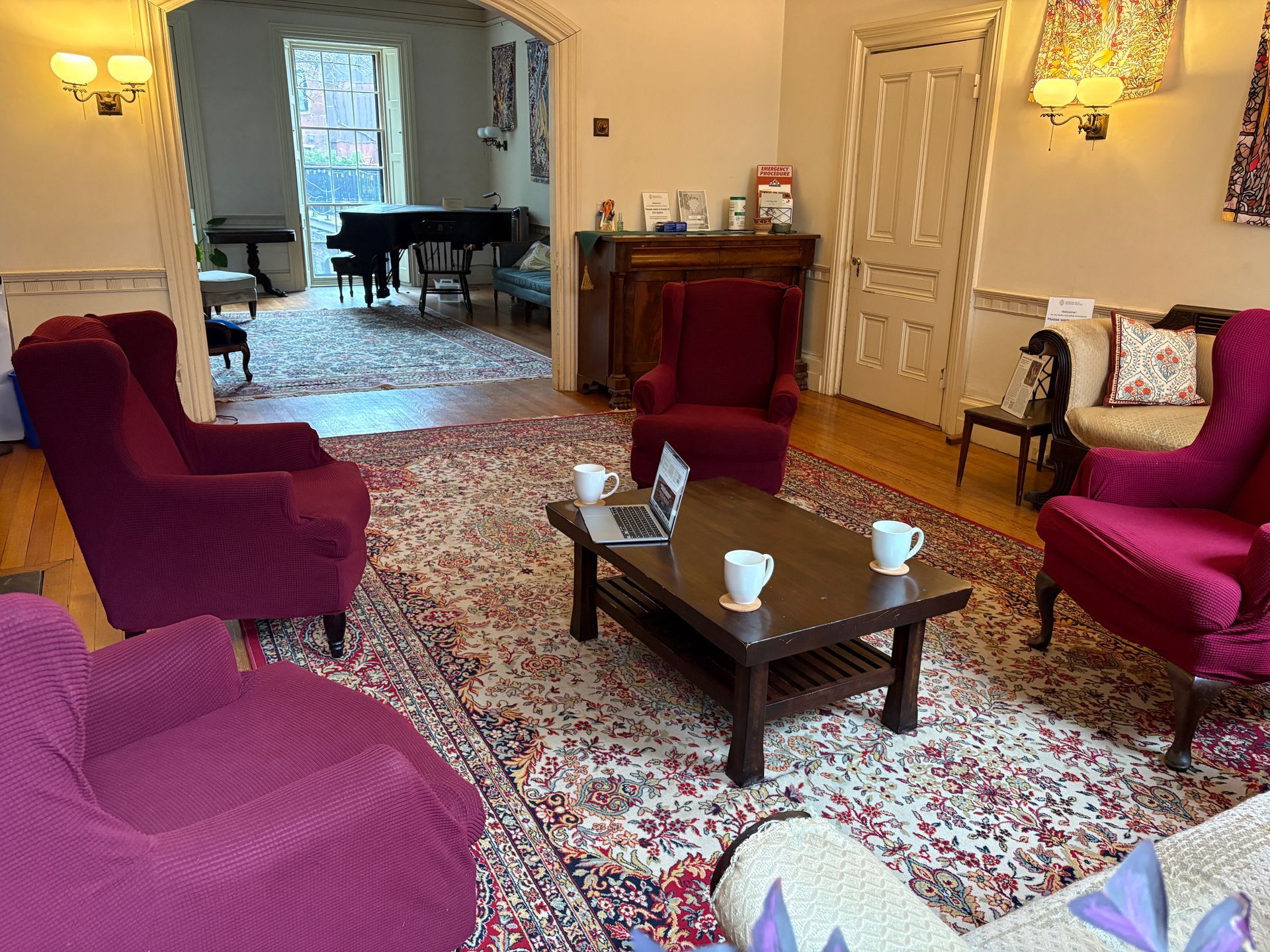 A cozy sitting area with maroon armchairs and a wood coffee table, opening into a dining room with a grand piano.