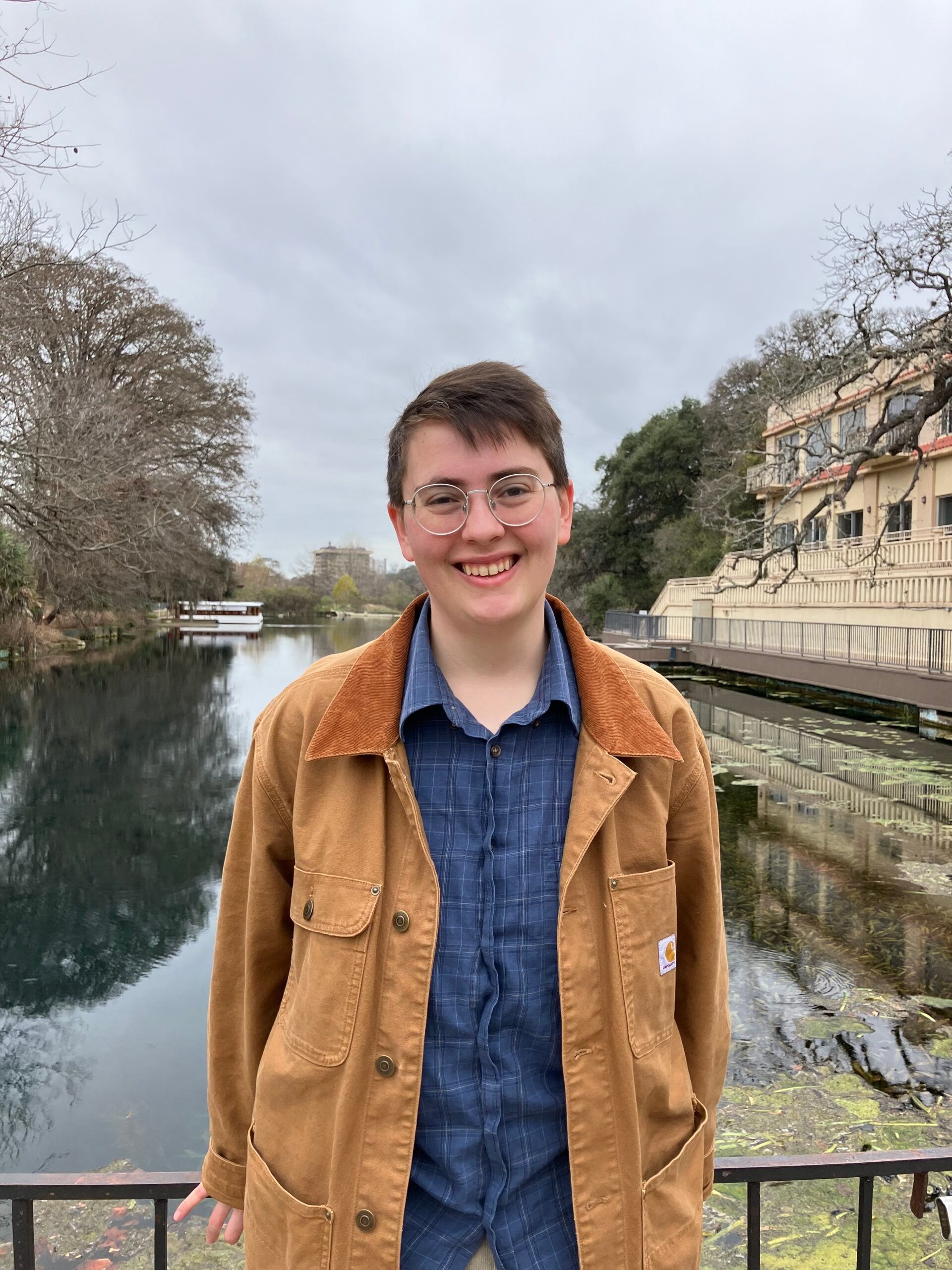 A person with glasses smiles while standing in front of a river and a building on an overcast day.