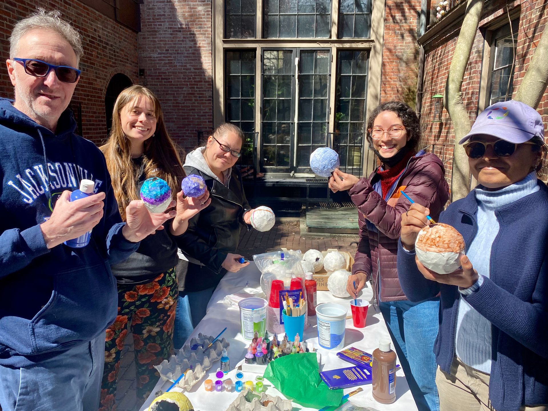 Five people stand outdoors around a table, painting small, spherical ornaments together in a garden setting.