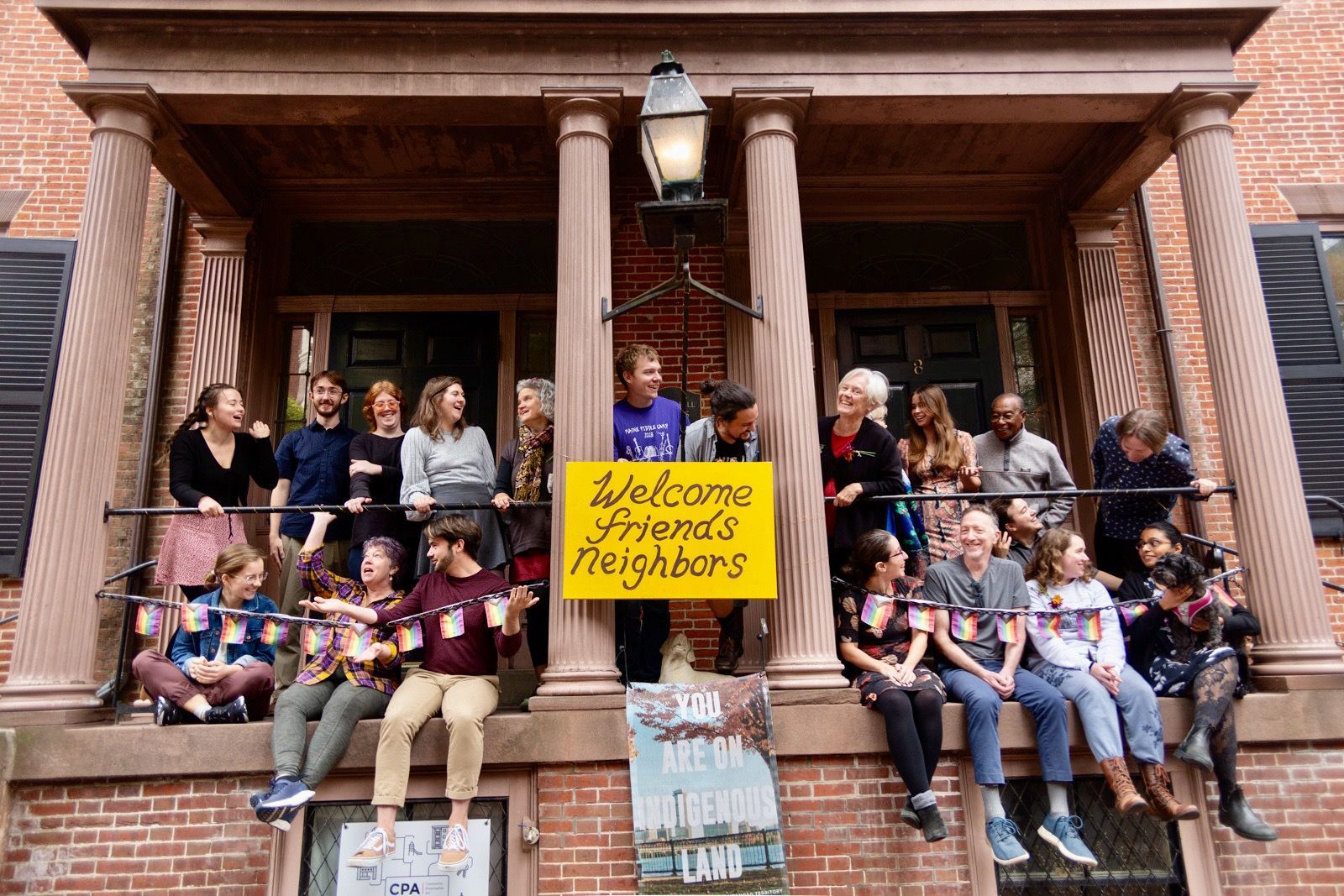 A diverse group sits on the front porch of a brick building, smiling behind a yellow sign that reads 