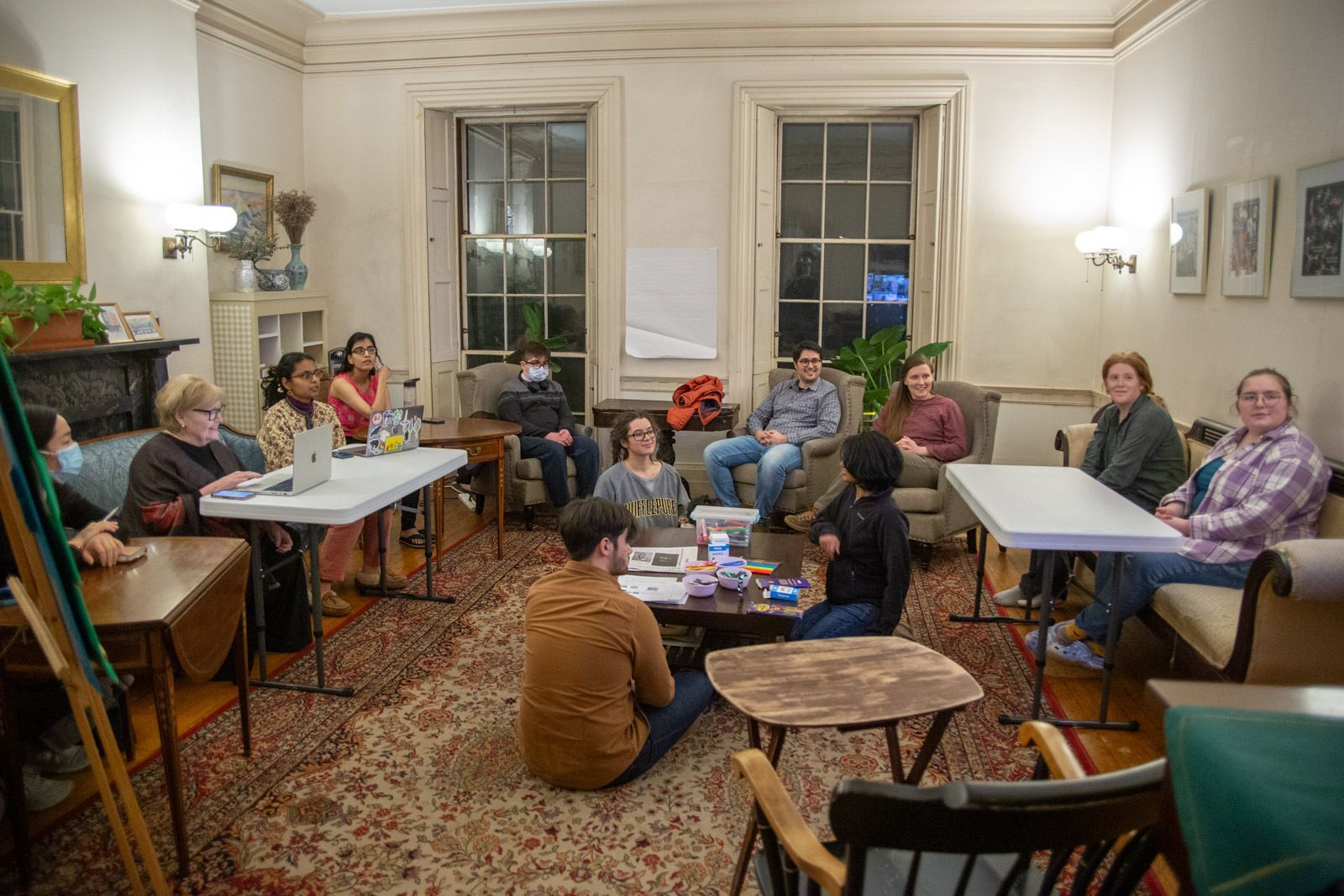A group of people sit around tables and in chairs in a softly lit, wood-paneled room for a meeting or study session.