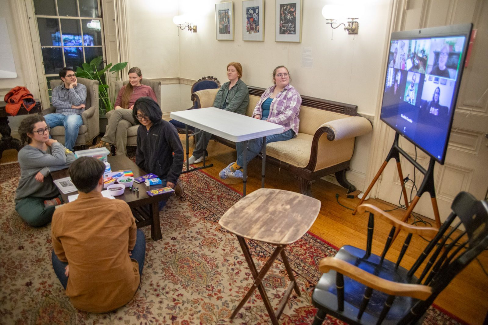 A group of people in a living room sit around a table and chair while viewing a large digital screen on a tripod.