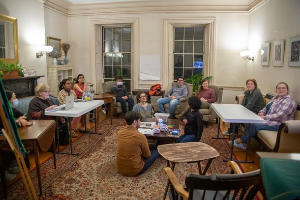 A group sits in a room with a patterned rug, tables, and windows for a discussion.