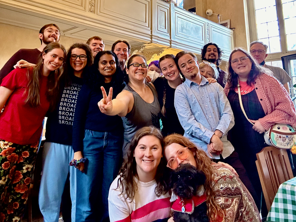 A group of people pose for a joyful photo in a brightly lit indoor space, with one person holding a small black dog.