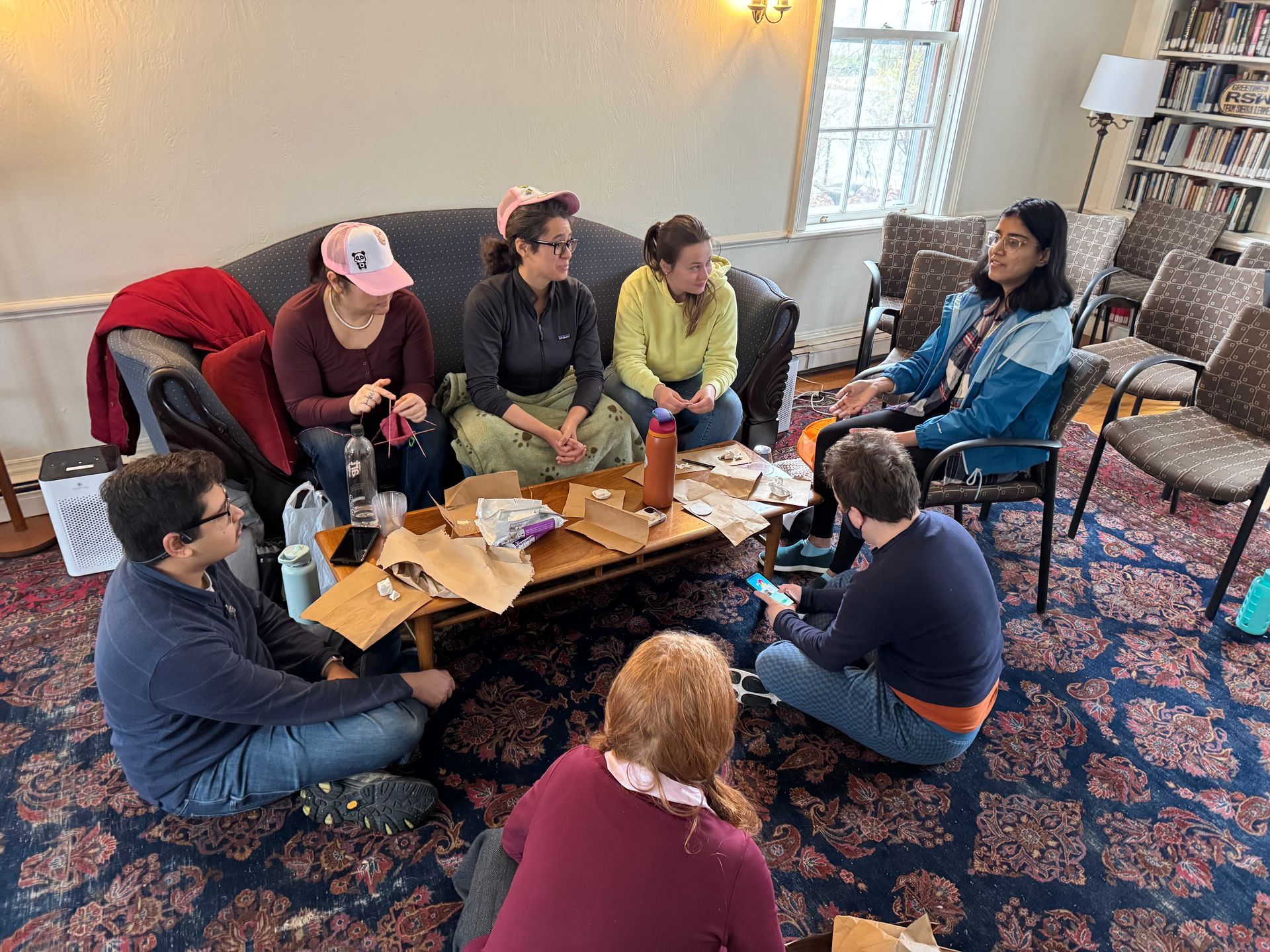 A group of people sit on a sofa and the floor in a room, working together on craft projects around a wooden coffee table.