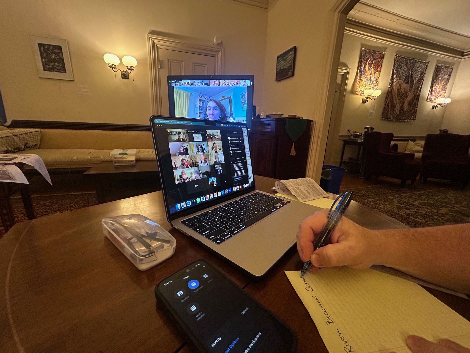 A person takes notes on a notepad during a video conference on a laptop, with a smartphone in the foreground.