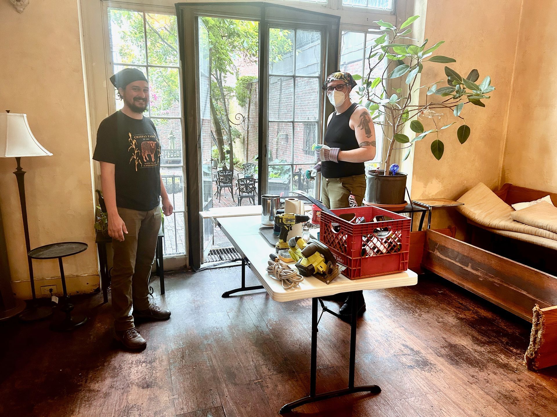 Two people stand on either side of a folding table holding a red crate and tools inside a room with a windowed door.