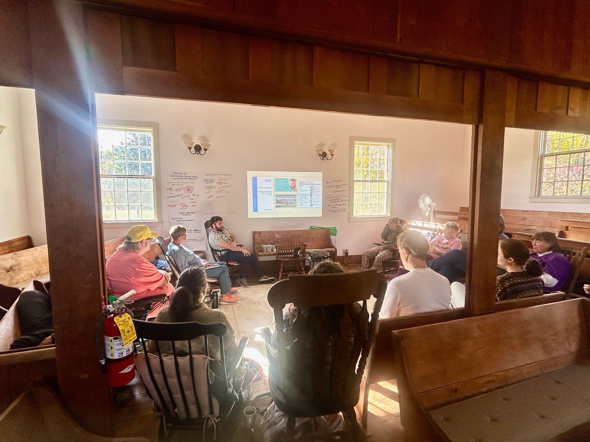 A group of people sits on benches and chairs in a wooden building, watching a presentation on a projected screen.
