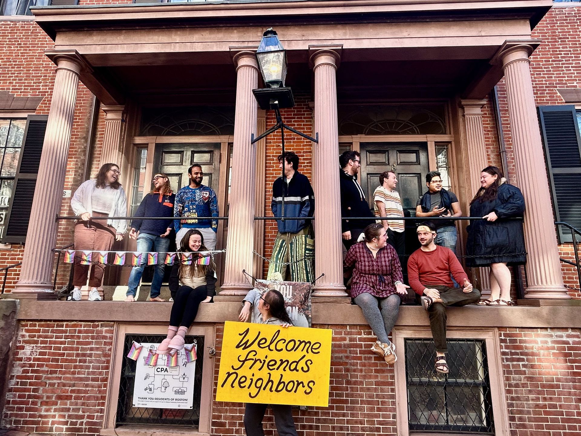 A group of people sit on the steps and porch of a brick building, holding a yellow sign that says 
