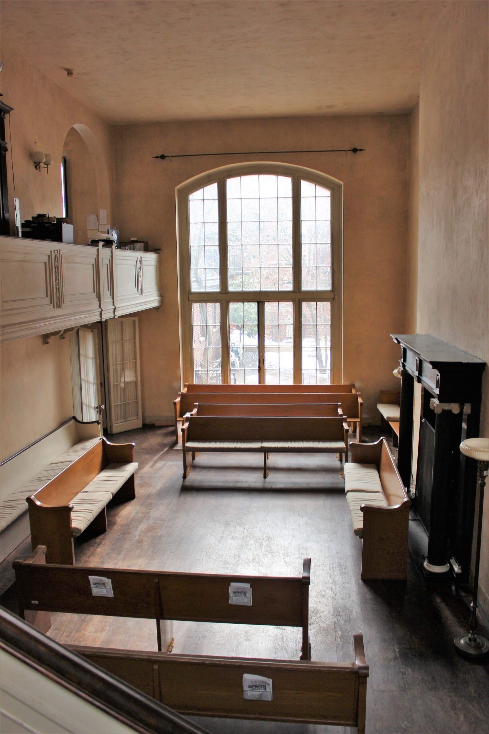 An interior view of a room with light brown walls, rows of wooden pews, a large arched window, and a dark fireplace mantel.