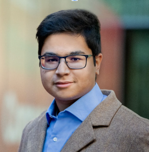 Headshot of a person wearing black-rimmed glasses, a blue collared shirt, and a brown textured blazer against a blurred wall.