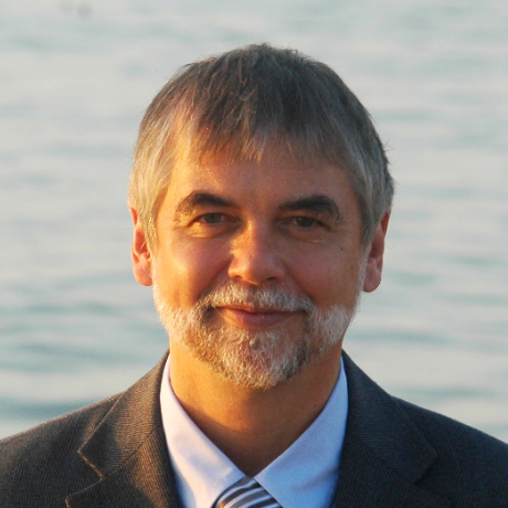 Portrait of a man with a gray beard wearing a suit and tie, smiling against a soft, blurred background of water.