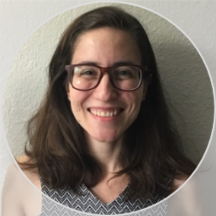 A smiling person with long dark hair and dark-rimmed glasses, wearing a patterned top, framed in a circle.