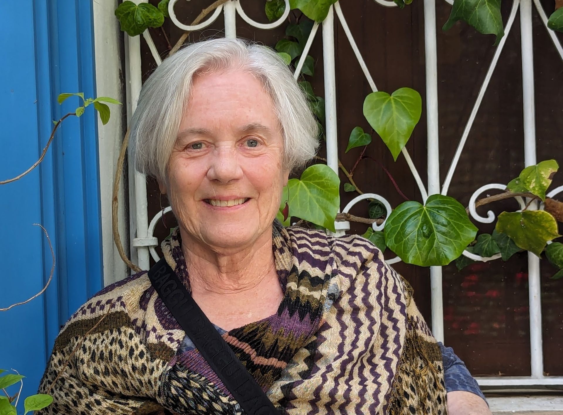 A smiling person with grey hair wearing a patterned shawl in front of a white metal grate with green ivy leaves.