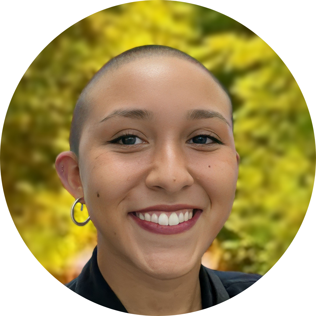 A smiling person with a shaved head and a gold hoop earring against a blurred, yellow-green foliage background.