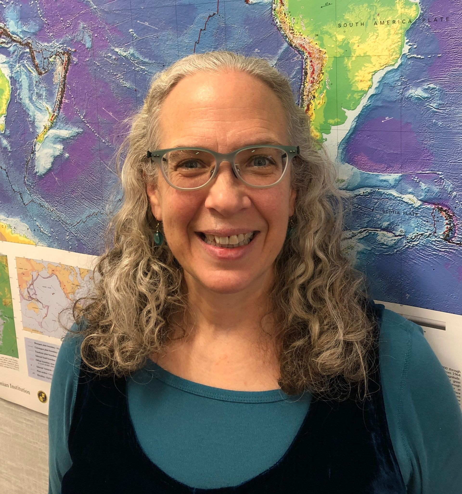 A smiling person wearing glasses and a teal shirt, standing in front of a colorful wall map of the ocean floor.