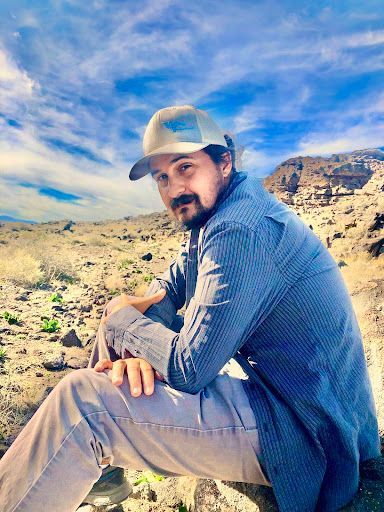A person wearing a cap and a blue button-down shirt sits in a desert landscape under a blue sky.