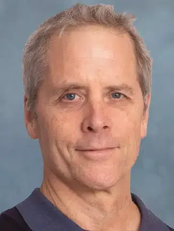 Head-and-shoulders portrait of a person with gray hair, wearing a dark blue shirt against a light blue background.