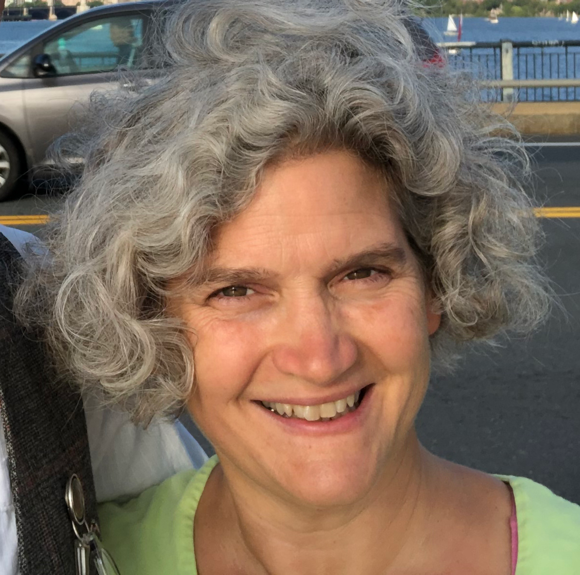 Smiling woman with wavy gray hair and a light green shirt, positioned outdoors near a body of water and a car.