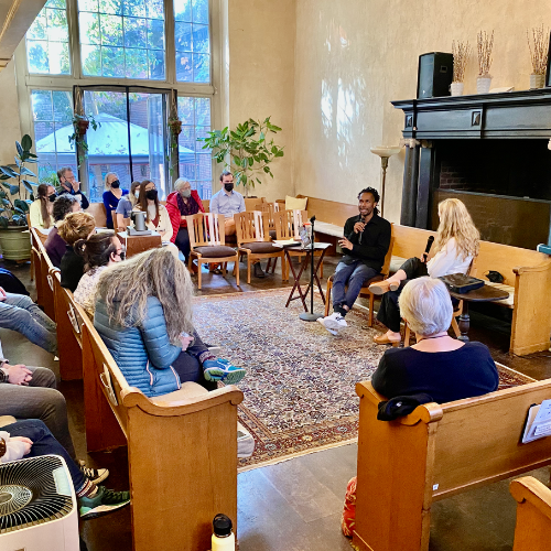 People in room, two speakers at table, seated audience in pews.  Window with natural light.