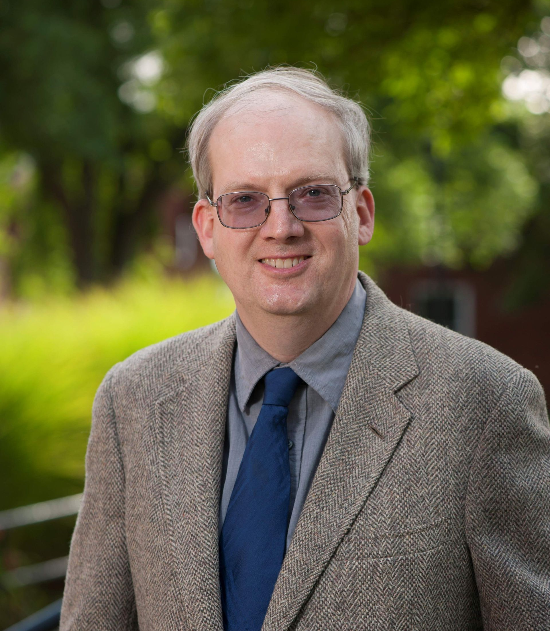 A person wearing glasses, a grey button-down shirt, a blue tie, and a textured blazer, standing outdoors.